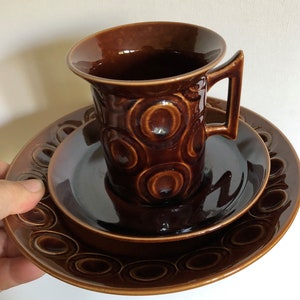 May include: A brown ceramic teacup and saucer set with a concentric circle pattern. The cup has a handle and the saucer has a raised rim.