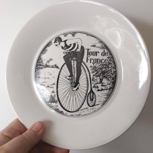 May include: White ceramic plate featuring a black and white illustration of a person riding a high-wheel bicycle. The plate has the words "Tour de France" printed on it. The plate is round and has a smooth, glossy finish.