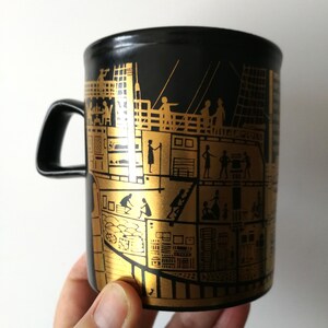 May include: Black ceramic mug with a gold design of a ship with people on board. The ship is depicted in cross-section, showing the interior of the ship with people in different rooms.