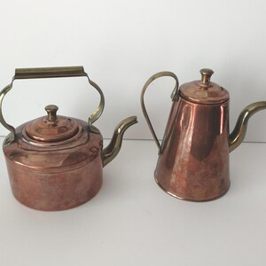 May include: Two copper teapots with brass handles and spouts. The teapot on the left has a rounded body and a hinged handle. The teapot on the right has a more angular body and a fixed handle. Both have lids.