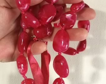 Peut inclure: Un long collier composé de grosses perles ovales roses. Le collier est noué avec un ruban rouge.