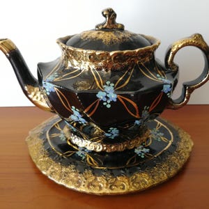 May include: Black teapot with gold accents and blue floral designs. The teapot has a matching saucer with a gold rim.