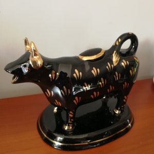 May include: Black ceramic creamer in the shape of a cow with gold accents. The creamer has a lid and a spout. It is sitting on a black base.