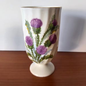 May include: A white porcelain vase with a fluted, pedestal base and a gold-trimmed rim. The exterior features a botanical illustration of purple thistle flowers with green, spiky leaves and stems painted against a soft, light blue background.