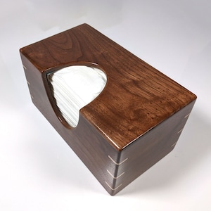 May include: A wooden tissue box holder made of dark brown walnut wood. The box has a cutout for the tissues and is finished with a clear coat.