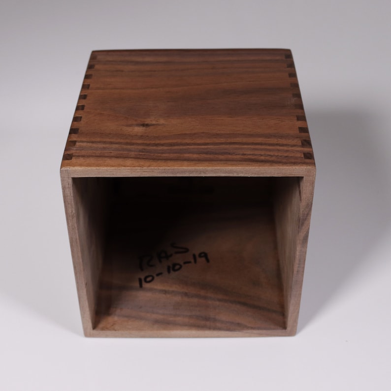 May include: A wooden box with a dark brown finish and a simple design. The box has a small opening on the top and is made of solid wood. The inside of the box is empty and has the inscription "RAS 10-10-19" written on the bottom.