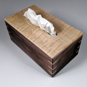 May include: A wooden tissue box cover with a light wood top and a dark wood base. The top has a natural wood grain pattern and a rectangular opening for tissues. The base has a smooth finish and is decorated with thin strips of wood.