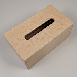 May include: A wooden tissue box cover made of light wood with a natural wood grain pattern. The box has a rectangular shape with a slot opening on the top for tissues.