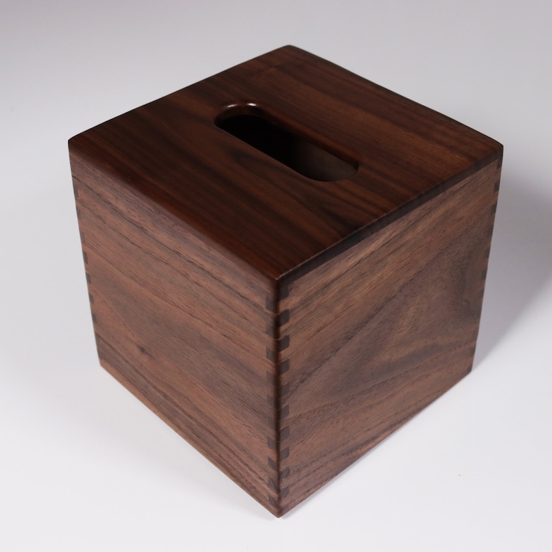 May include: A dark brown wooden tissue box cover with a rectangular opening for tissues. The box is made of solid wood and has a smooth finish.