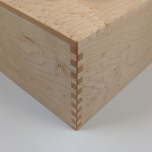 May include: Close-up of a wooden box with a light wood finish. The box has a mitered corner with a visible dovetail joint.