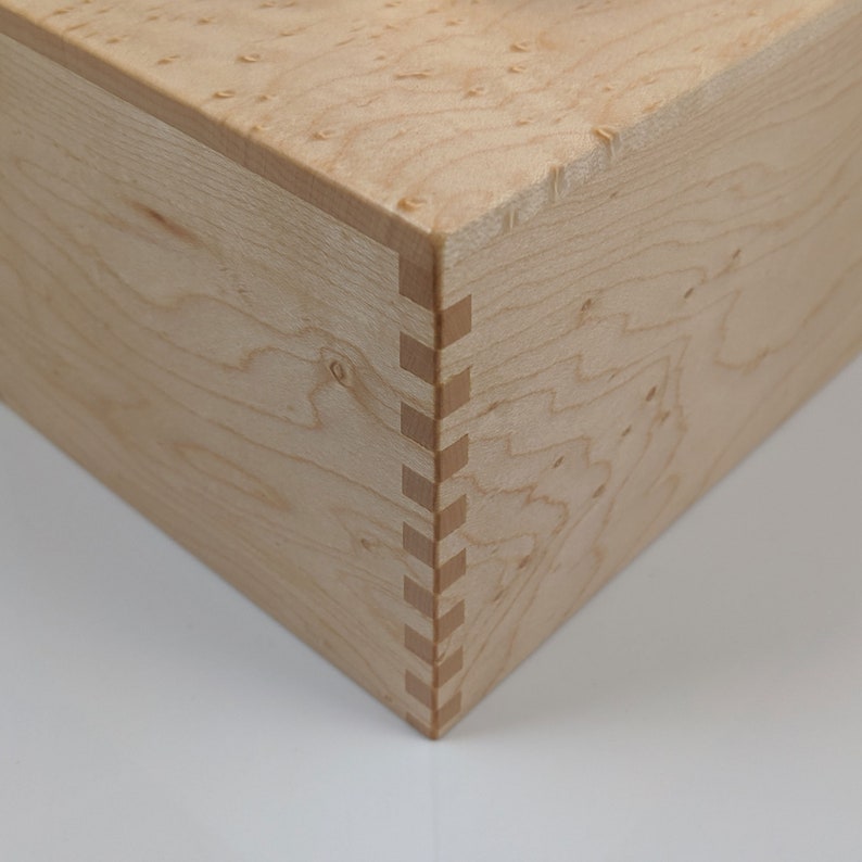 May include: Close-up of a wooden box with a light wood finish. The box has a mitered corner with a visible dovetail joint.
