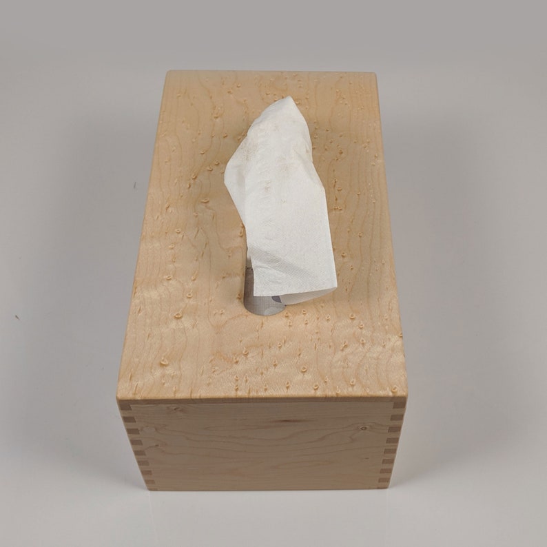 May include: A wooden tissue box cover with a natural wood finish and a visible wood grain pattern. The box has a rectangular shape and a slot for tissues to be pulled out.