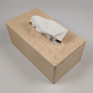 May include: A wooden tissue box cover made of maple wood with a natural wood finish. The box has a slot for pulling out tissues.