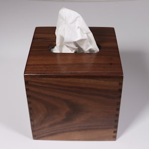 May include: A square wooden tissue box holder made of dark walnut wood. The box has a slot for tissues and a lid.