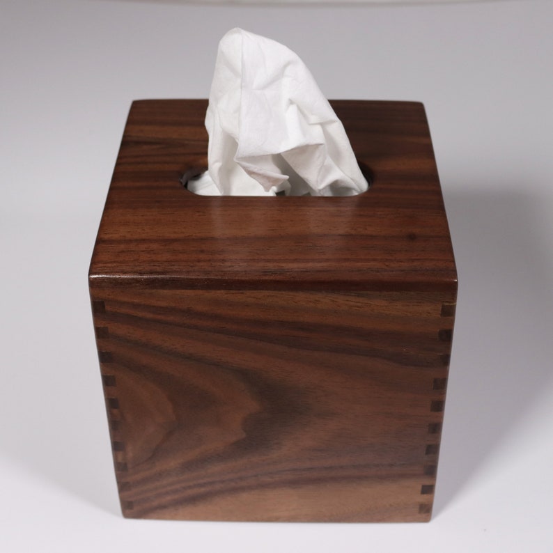 May include: A square wooden tissue box holder made of dark walnut wood. The box has a slot for tissues and a lid.
