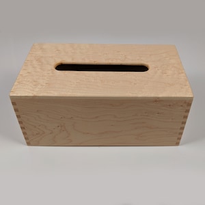 May include: A wooden tissue box cover with a natural wood finish and a rectangular opening for tissues. The box is made of light wood with a grain pattern.