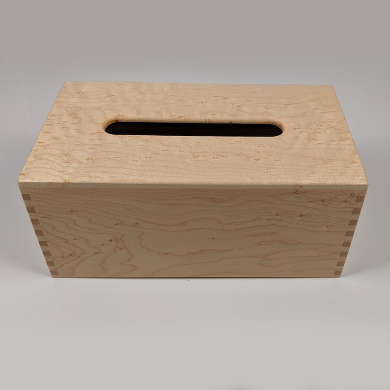 May include: A wooden tissue box cover with a natural wood finish and a rectangular opening for tissues. The box is made of light wood with a grain pattern.