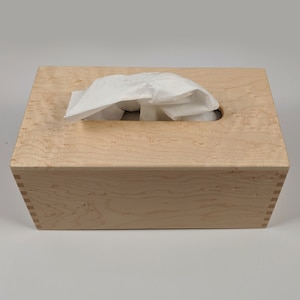 May include: A wooden tissue box cover made of maple wood with a natural finish. The box has a rectangular shape and a slot for pulling out tissues.