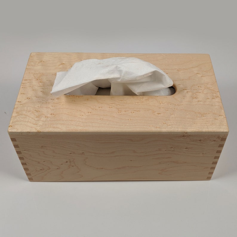 May include: A wooden tissue box cover made of maple wood with a natural finish. The box has a rectangular shape and a slot for pulling out tissues.