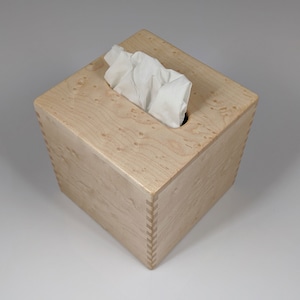 May include: A square wooden tissue box cover made of light wood with a natural wood grain pattern. The box has a slot for tissues and is made with a dovetail joint.