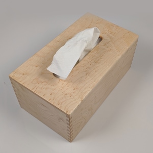 May include: A wooden tissue box cover made of maple wood with a natural wood finish. The box has a rectangular shape and a slot for tissues to be pulled out.