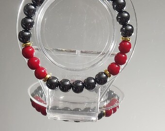 May include: A beaded bracelet featuring alternating red and black beads. The bracelet has gold-colored spacers between the red beads. The bracelet is displayed on a clear, circular stand.