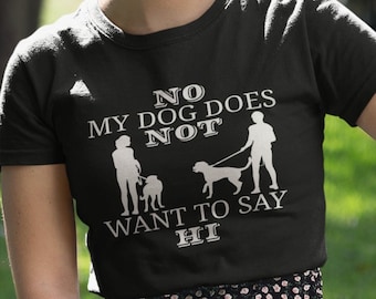 May include: A woman wearing a dark green t-shirt with the text "NO MY DOG DOES NOT WANT TO SAY HI" in white. She is holding a pink leash and is kneeling in a grassy area with a white dog.