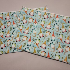 May include: Three zippered pouches with a light blue background and a repeating pattern of gnomes wearing colorful hats and holding lanterns. The gnomes are surrounded by green pine branches and white snowflakes.