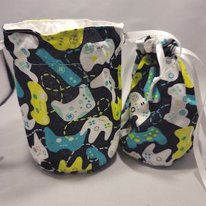 May include: Two drawstring pouches with a black background and a pattern of white, teal, and yellow video game controllers. The larger pouch is cylindrical, and the smaller one is rounded. Both have white ribbon drawstrings.