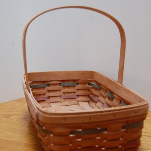 May include: A woven basket with a natural wood finish and a handle. The basket has a rectangular shape and is made of woven strips of wood. The basket has a light brown color and is empty.