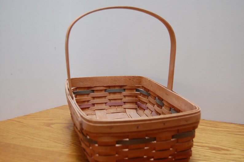 May include: A woven basket with a natural wood finish and a handle. The basket has a rectangular shape and is made of woven strips of wood. The basket has a light brown color and is empty.