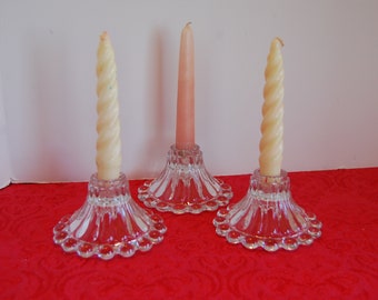 Peut inclure: Trois bougeoirs en verre avec un bord festonné et un motif cannelé. Deux bougies sont blanches et une est rose.