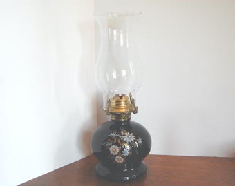 Peut inclure: Une lampe à huile vintage avec une base noire et une cheminée en verre transparent. La base est ornée d'un motif floral blanc, doré et bleu. La lampe a un raccord en métal couleur laiton et repose sur une surface en bois.