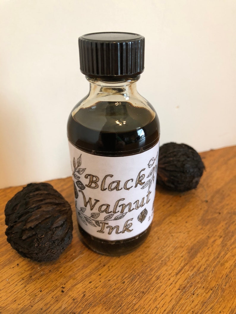 May include: A clear glass bottle of Black Walnut Ink with a black cap and a white label. The label has the words "Black Walnut Ink" in a decorative font. Two black walnuts are next to the bottle on a wooden surface.
