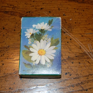 May include: A small, rectangular box with a blue background and a floral design featuring white daisies with yellow centers. The box has a green border and is resting on a wooden surface.