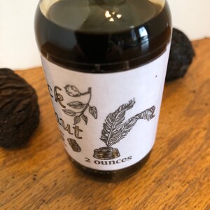 May include: A clear glass bottle filled with a dark liquid, labeled "Black Walnut" with botanical illustrations and a quill pen. The label indicates a 2-ounce volume. Two black walnuts are visible on a wooden surface.