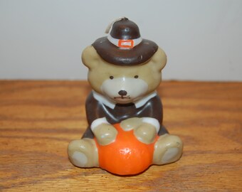 May include: A Thanksgiving-themed candle shaped like a teddy bear. The bear wears a brown pilgrim hat with a white and orange accent, a brown coat, and a white collar. It holds an orange pumpkin. The candle is on a wooden surface.