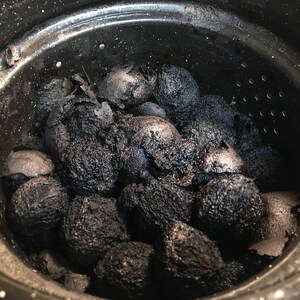 May include: A close-up shot of a black pot filled with charred, oval-shaped objects. The objects are dark, with a rough, textured surface, and appear to be partially burned. The pot is black with a silver rim and small holes near the top.