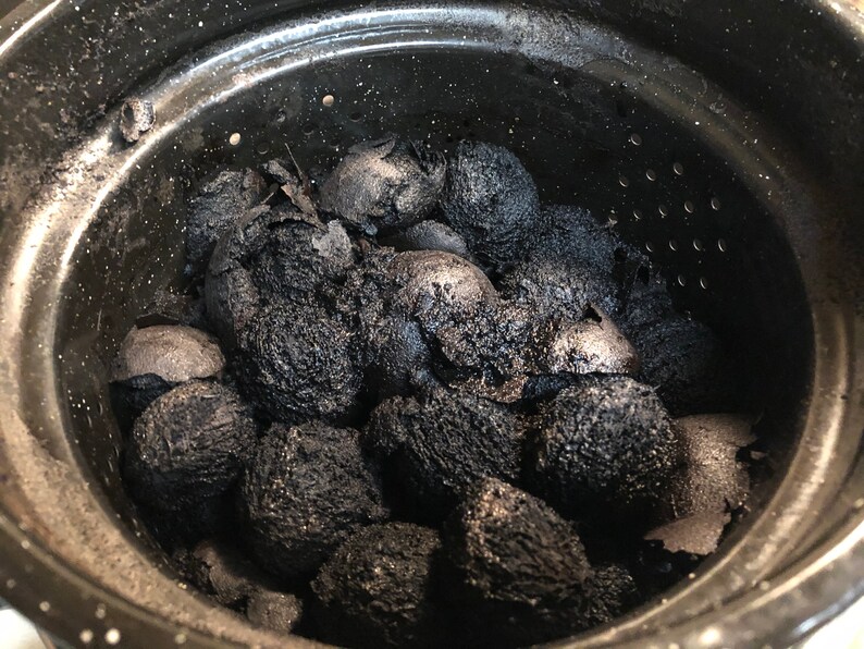 May include: A close-up shot of a black pot filled with charred, oval-shaped objects. The objects are dark, with a rough, textured surface, and appear to be partially burned. The pot is black with a silver rim and small holes near the top.