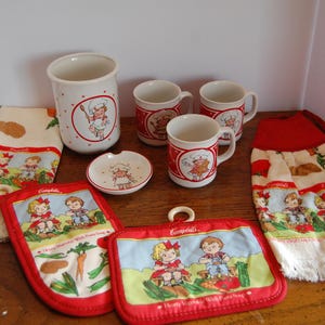 May include: A collection of vintage Campbell's kitchenware featuring a chef-themed design. Includes mugs, a utensil holder, a small dish, pot holders, and dish towels. The items are primarily white with red accents and illustrations of children.