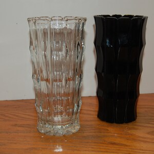 May include: Two glass vases, one clear and one black, with a textured, ribbed design. The vases are sitting on a wooden surface.