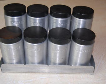 Peut inclure: Un ensemble de huit pots à épices en argent avec des couvercles noirs. Les pots sont disposés en rangée sur un support en argent. Les pots sont étiquetés avec les noms de différentes épices, notamment "Sel", "Poivre", "Ail", "Oignon", "Origan", "Basilic", "Thym" et "Romarin".