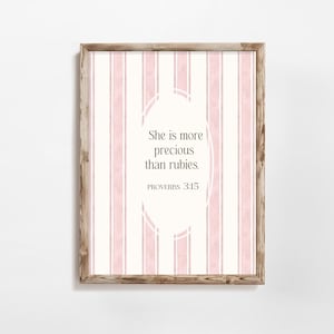 Może przedstawiać: Oprawiony wydruk z jasną drewnianą ramą. Grafika przedstawia różowo-białe paski w tle z tekstem "She is more precious than rubies. PROVERBS 3:15" w białym owalu.
