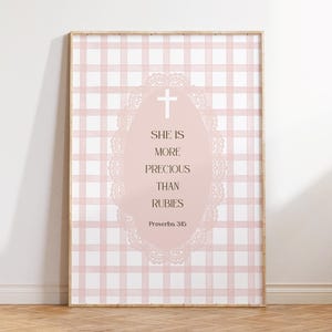 Pode incluir: Uma impressão emoldurada com um fundo de padrão xadrez rosa e branco. A impressão tem um design oval central com uma cruz e o texto "SHE IS MORE PRECIOUS THAN RUBIES Proverbs 3:15". A moldura é de cor de madeira clara.