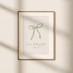 May include: Framed art print featuring a watercolor illustration of a light green bow. The text "I am a child of God, John 1:12" is written below the bow in a delicate script. The frame is a warm brown color.