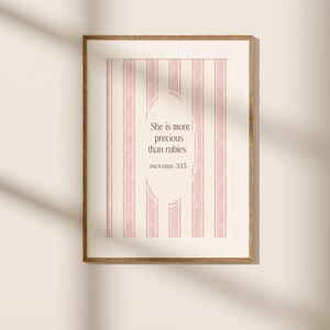 May include: A framed print featuring a vertical striped background in cream and pink. The print includes the text "She is more precious than rubies. PROVERBS 3:15" in a decorative font, set within an oval shape.