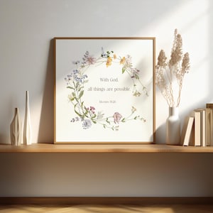 May include: Square framed print featuring a floral wreath with text that reads "With God, all things are possible. Matthew 19:26." The print is in a wooden frame and sits on a wooden shelf. A white vase with dried flowers is to the right.