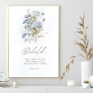 Puede incluir: Una impresión artística enmarcada con un arreglo floral en acuarela en tonos suaves de azul, morado y verde. El texto "Behold, The Lamb of God, who takes away the sin of he world. John 1:29" se muestra en una elegante escritura.