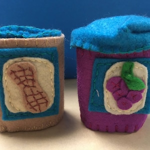 May include: Two felt jars, one with a peanut design and the other with a grape design. The peanut jar is brown with a blue lid and the grape jar is purple with a blue lid.
