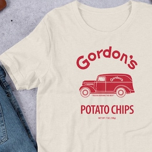 Puede incluir: Una camiseta beige con un gráfico rojo de un camión de reparto vintage con el texto "Gordon's" y "Potato Chips" impreso en él. El camión tiene el texto "Gordon's" en el lateral y las palabras "Trucks Serving the Best" impresas debajo. El texto "Net Wt 7 oz (198g)" está impreso debajo de las palabras "Potato Chips".
